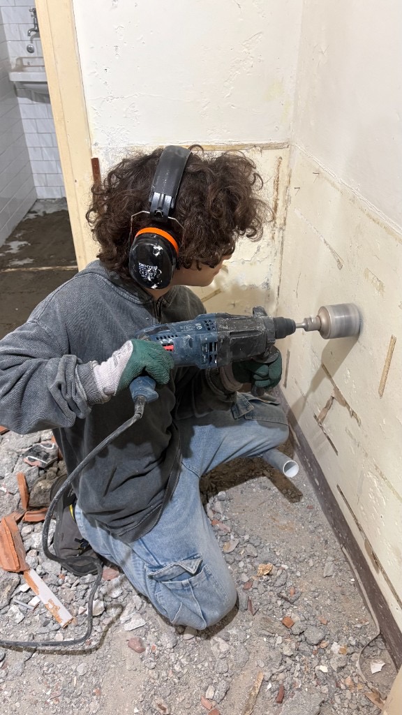 Partecipante al laboratorio di autocostruzione che esegue un foro a carotiere su un muro interno, con cuffie e guanti di protezione