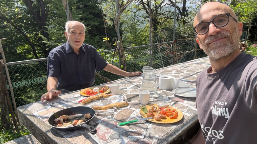 Due persone a pranzo all'aperto a un tavolo di pietra con mosaico, tra il verde